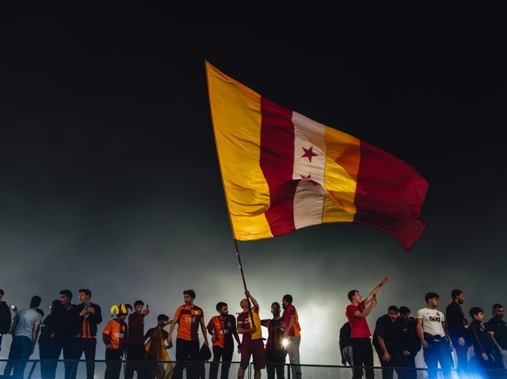 Anhänger schwenken eine riesige gelb-rote Vereinsfahne vor einem dunklen Nachthimmel. Spüren Sie die leidenschaftliche Stimmung bei einer Fußballreise in die Türkei.
